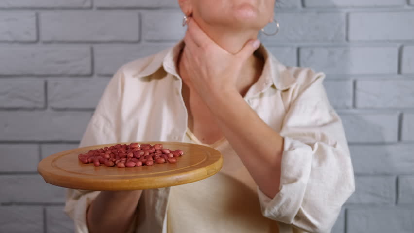 Woman with a peanut allergy touching her throat. Female individual suffering from an allergic reaction, holding a wooden plate with peanuts while touching her throat in distress due to swelling