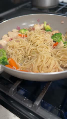 Vertical close-up of lo mein noodles stir fried with chicken and vegetables in a skillet, being mixed while cooking on a stovetop.
