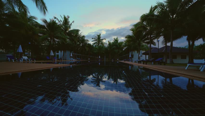 Watch as the dawn light changes over a hotel resort in Thailand. The pool reflects the sky while palm trees stand tall. This shows the start of a new day in a tropical setting.
