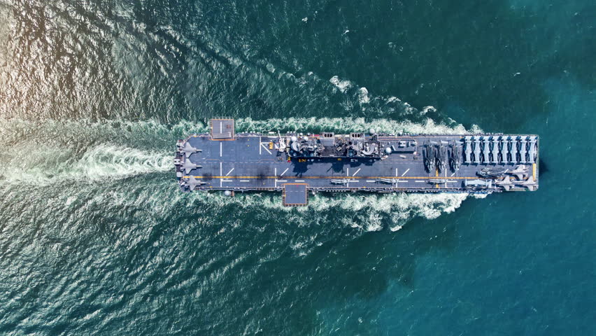 nuclear navy aircraft carrier sailing in the deep blue ocean. Cinematic shot of a military warship deck loaded with fighter jets leaving a white wake.