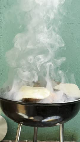 Meat and tortillas cooking on outdoor barbecue grill with thick white smoke rising. Street food and backyard grilling atmosphere, close-up cooking process.