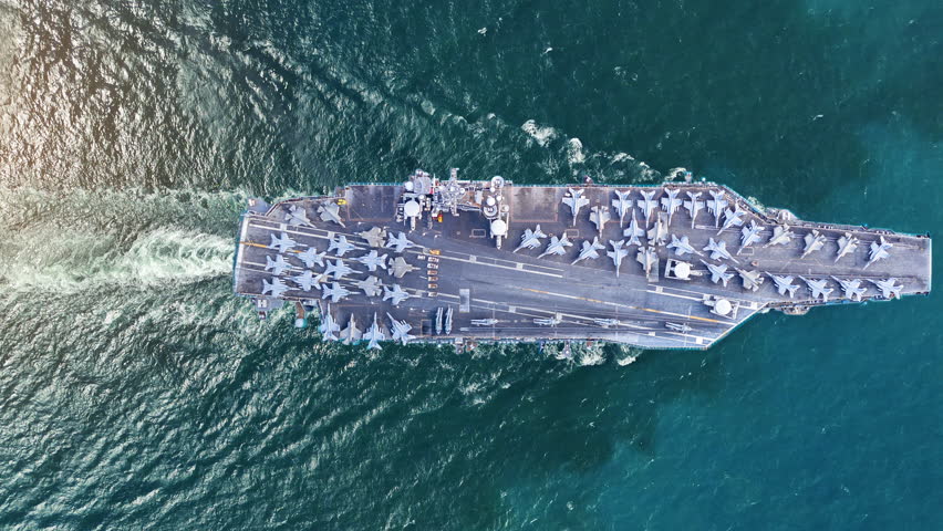 nuclear navy aircraft carrier sailing in the deep blue ocean. Cinematic shot of a military warship deck loaded with fighter jets leaving a white wake.