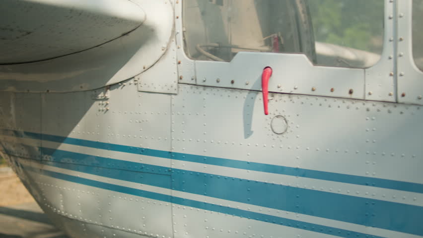 A smooth cinematic gimbal shot moving along the white riveted aluminum fuselage of a vintage military observation aircraft. The shot highlights the blue horizontal stripes, weathered skin texture, and a red door latch handle.