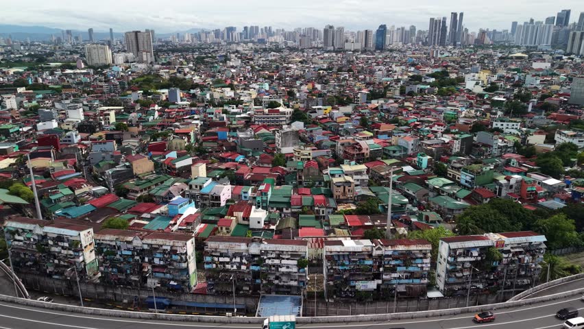 High-angle wide shot of dense old residential blocks along a multi-lane expressway, with modern high-rise towers in the background in Manila, Philippines under cloudy skies.