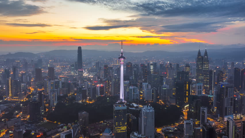 Golden sunrise time lapse view of overlooking urban forest and roads with KL skyline in background in Asia. Kuala Lumpur, Malaysia. The scene features world-famous architectural icons including the Petronas Twin Towers, Menara Kuala Lumpur, and Merdeka 118 rising above a dense urban landscape.. Dolly Zoom motion timelapse.