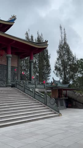 Traditional Chinese-style temple architecture featuring a multi-tiered teal roof with ornate dragon sculptures and bright red pillars. The building sits on a stone terrace overlooking a misty mountain landscape. Cultural and religious landmark photography.