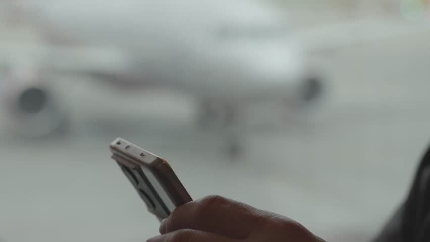 A person holds a smartphone while waiting at the airport for their flight to board. Planes are parked at the terminal in the background.