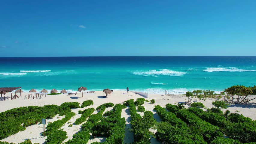 View of vibrant turquoise water waves hitting the shore at a beach with straw huts, beach chairs, and green bushes against a bright blue sky on a sunny day.