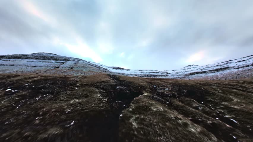 FPV style low altitude aerial shot moving rapidly over a muddy and mossy mountain path. The ground is textured with grass and light snow, leading up to a ridge under a cloudy sky.