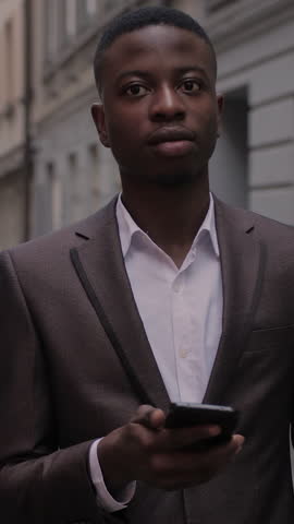 Handsome african guy in stylish business suit checking emails on modern smartphone while standing on city street. Concept of work, success and technology.