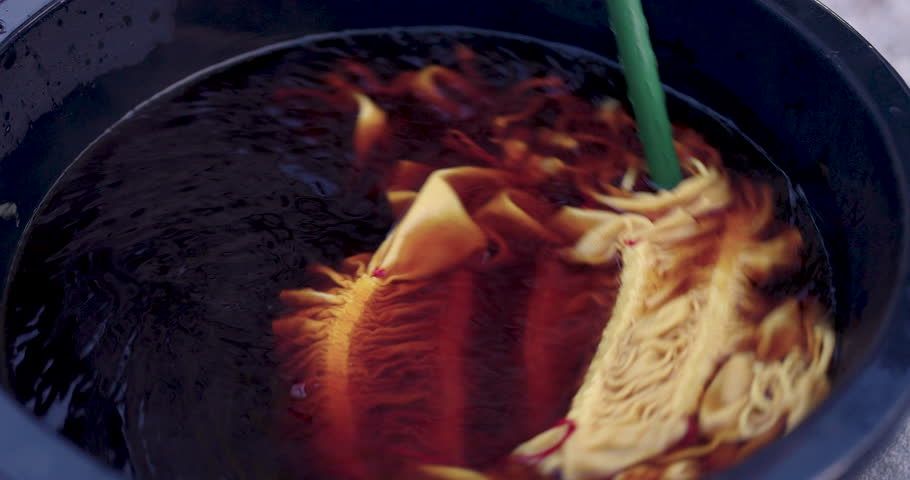 Macro view shows indigo dye whirlpool as senior Asian woman artisan stirs shibori cloth, ripples pull fabric inward, color spreads through cotton, resist lines stay bright, motion in basin, outdoor.