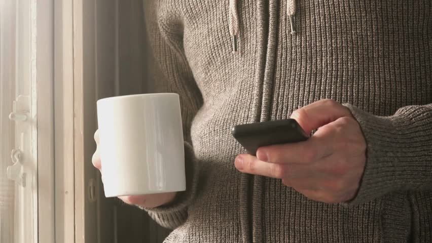 Person in sweater near bright window texts on phone while holding white mug. Casual indoor daylight scene. Relaxed mood and morning routine in a cozy home.