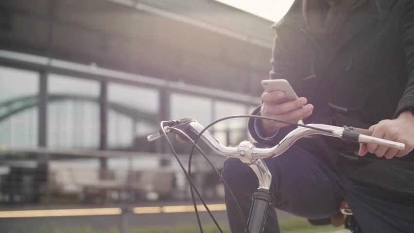 Person on bicycle uses cell phone outdoors by architecture mirroring bridge. Commuter checks smartphone in bright sunlight near building in Cologne, Germany.