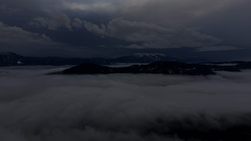 Heavy cloud layers drift slowly over remote highlands and fog-filled valleys in a dark atmospheric landscape.