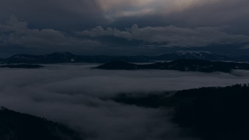 Heavy storm clouds move slowly over quiet hills and fog-filled valleys creating a dark and atmospheric natural scene.