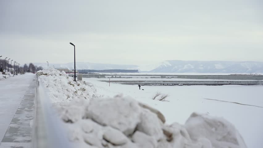 Snowy winter landscape featuring a person walking with a dog along a frozen river, viewed from an embankment with piles of snow against an overcast sky and distant mountains