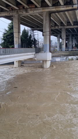 Raging muddy river flooding with powerful currents flowing under concrete bridge supports during a severe storm, highlighting climate change and natural disaster damage