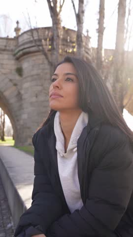 Serene woman with long hair enjoying a sunny day sitting by a stone bridge in a city park