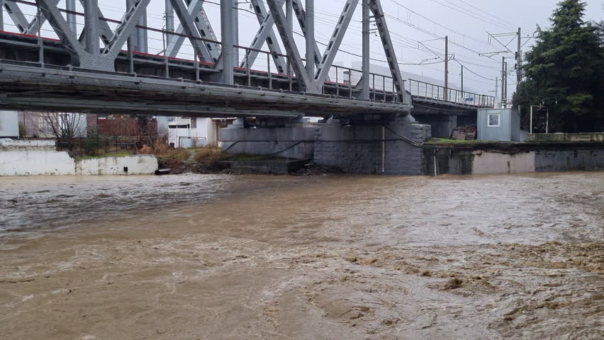 Raging muddy river flowing swiftly under a railway bridge during a flood, illustrating the destructive power of a natural disaster and extreme weather conditions
