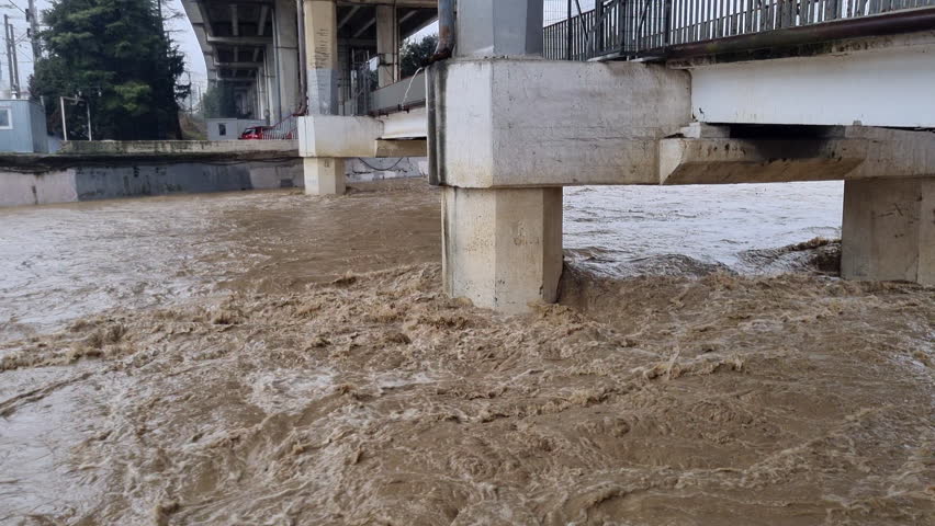 Raging muddy river overflowing its banks, powerfully flowing under a concrete bridge during a severe weather event, showcasing the force of a natural disaster
