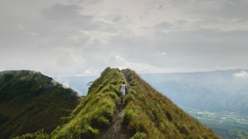 Man running narrow mountain ridge with a scenic landscape view. Ascending a trail as an active volcano emits steam into the cloudy sky. Representing endurance. Freedom. And exploration in nature