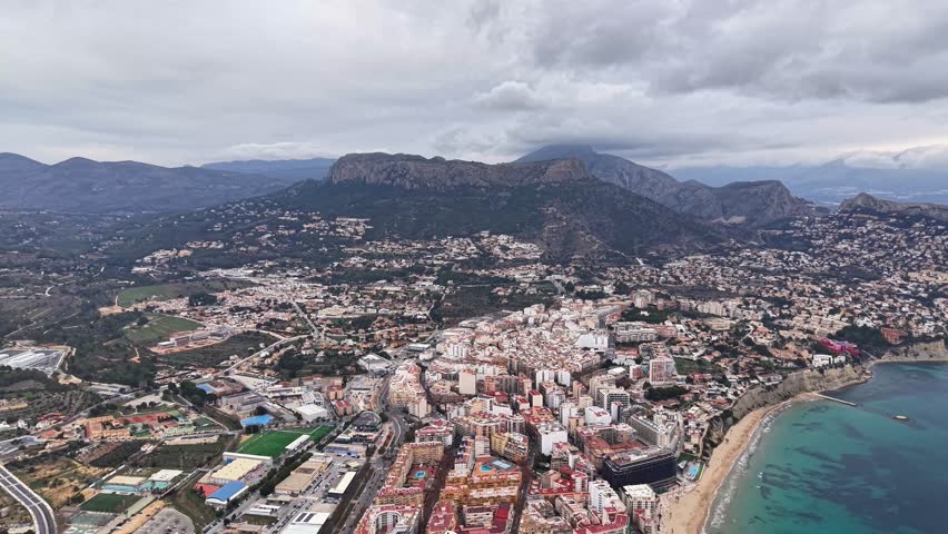 Aerial Hillside City Under Low Clouds, Dense Urban Blocks Meeting Sandy Bay, Mountain Rim In Background,