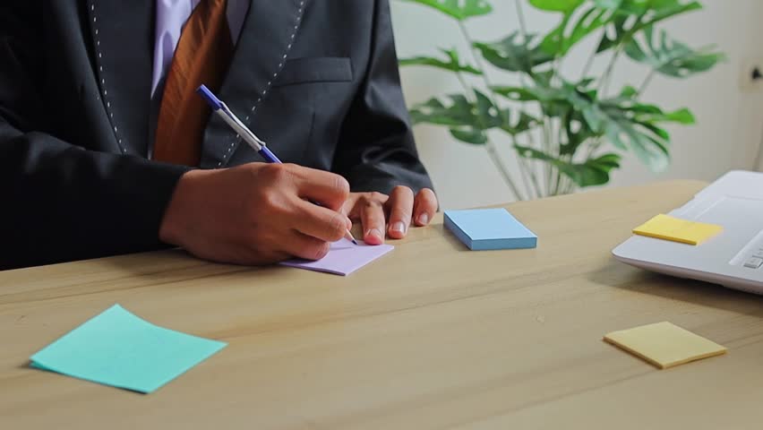 Professional businessman in black suit writes notes on purple sticky notes at wooden desk