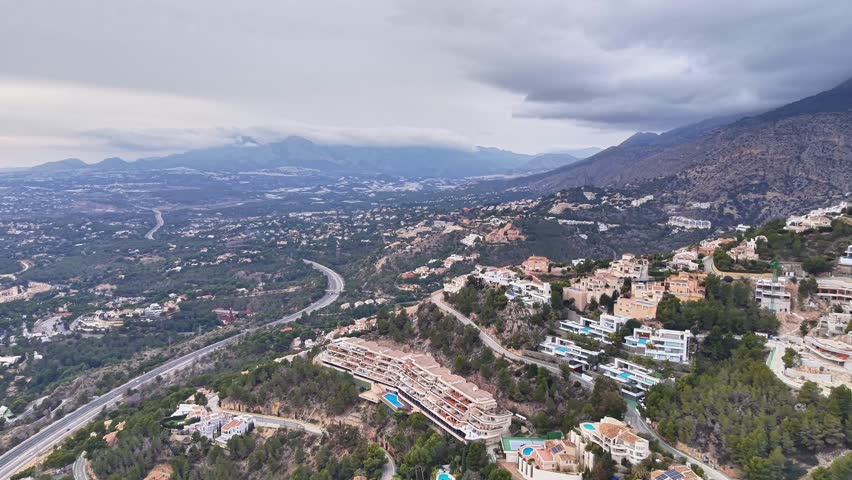 moody hillside residential aerial scene with clustered villas, pools, winding highway and overcast sky