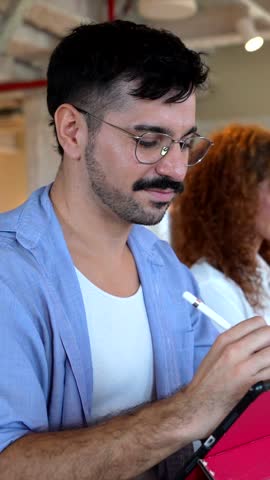 Young handsome graphic designer with a mustache using a stylus on a digital tablet, working alongside diverse colleagues in a modern, collaborative open plan office or coworking space