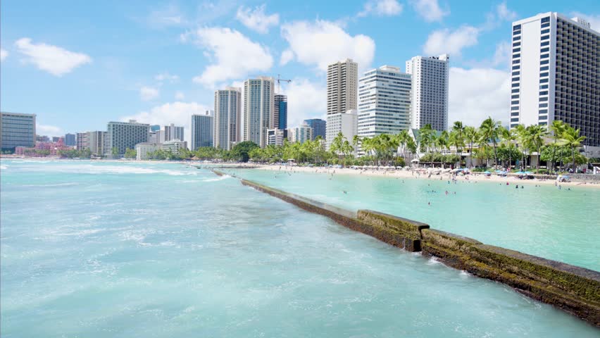 Beautiful panoramic view of the famous waikiki beach in honolulu, hawaii, showing the city skyline, hotels, and turquoise ocean waves washing over a stone breakwater on a sunny summer day