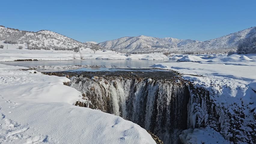 A powerful waterfall flows through a snowy winter landscape, surrounded by forested hills and bright blue sky, highlighting the contrast between moving water and frozen terrain.  
📍Marivan, Kurdistan 