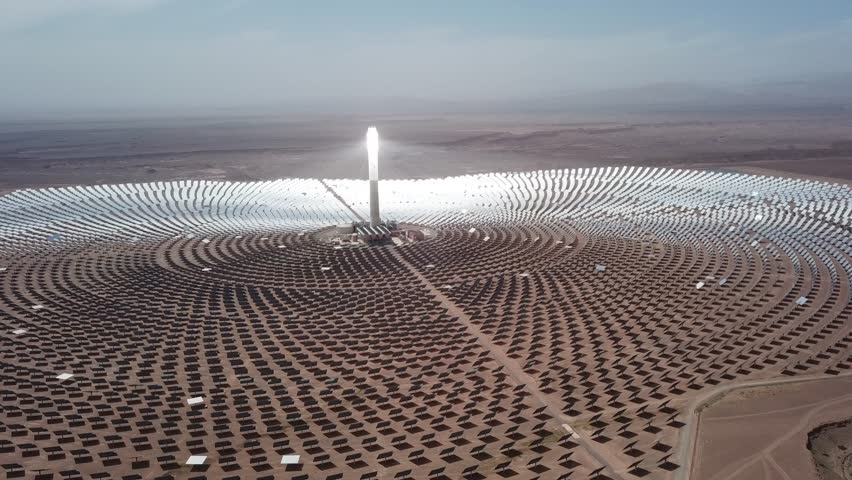 Aerial drone shot of the Noor III Concentrated Solar Power (CSP) plant. Features the glowing central tower surrounded by thousands of heliostat mirrors in the Moroccan desert.
