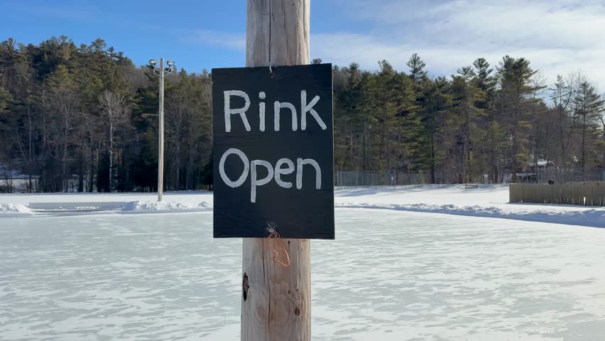 Outdoor ice rink in winter with a wooden pole sign reading “Rink Open,” surrounded by snow and trees.