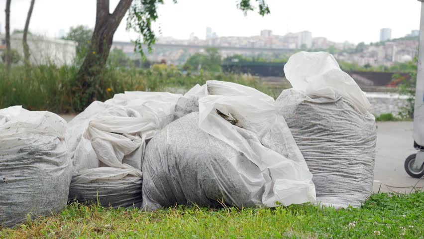 Pile of large plastic garbage bags filled with debris on green grass along city park road