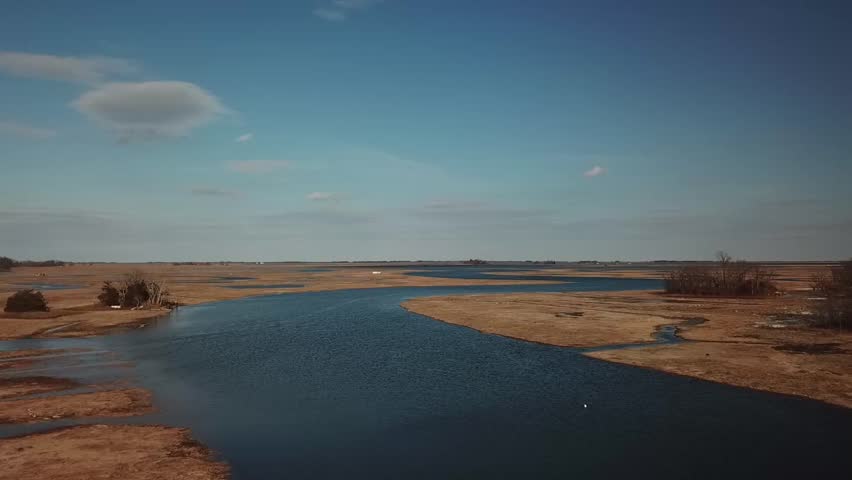 Wide aerial view of Essex Bay salt marsh in Massachusetts, winding tidal channel through golden marsh grass under blue sky, calm water and vast coastal wetlands, daytime.