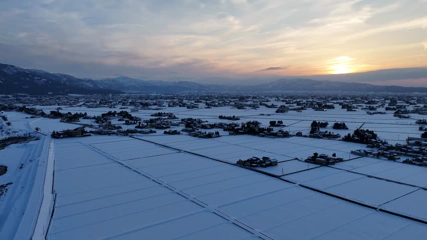 A snowy landscape with a beautiful sunset in the background. The sky is filled with clouds, and the sun is setting, casting a warm glow over the scene