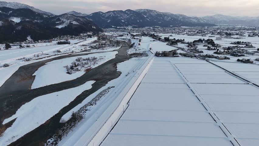 A snowy landscape with a river and mountains in the background. The sky is cloudy and the snow is covering the ground