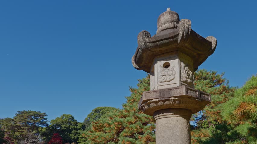 A low-angle shot of a classic Japanese stone lantern (toro) against a vivid blue sky, surrounded by lush pine trees and autumn foliage in the historic Rikugien Garden.