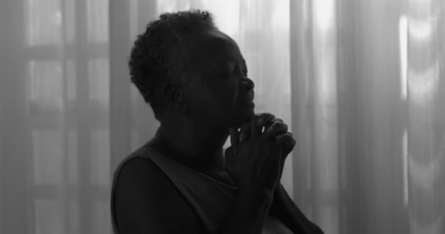 Elderly African American woman praying softly by window in monochrome, eyes closed, enduring faith, quiet hope, spiritual perseverance in later life