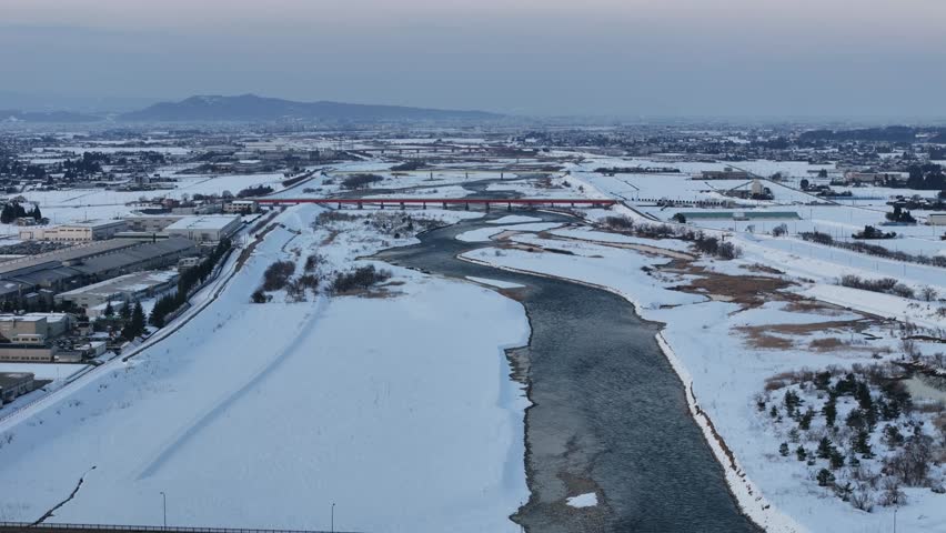 A snowy landscape with a river and a bridge. The sky is cloudy and the snow is covering the ground
