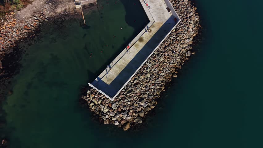 People are walking on a pier made of rocks by the sea. The water is calm, and some are fishing. It is sunny, and the scene shows a beach area with no clouds.