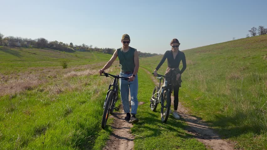 Two people ride bicycles on a scenic outdoor path surrounded by trees and blue sky, enjoying the fresh air.