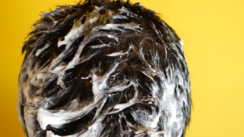 Rear view of young man washing his dark hair with white soapy foam against yellow background