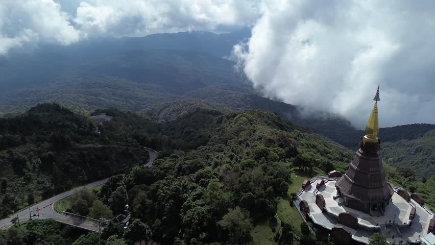 Golden pagoda standing on a mountain summit, surrounded by lush green forest and winding roads under dramatic cloudy skies.