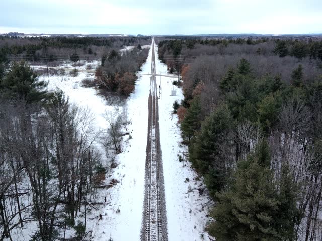 Aerial drone footage moving forward along railroad tracks near a road intersection in a snowy winter landscape.