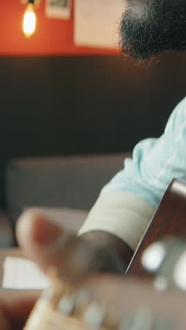Vertical tilt up shot of African American man wearing headphones performing guitar chords and vocalizing into microphone in home recording studio