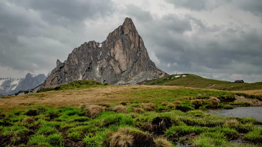 A majestic jagged mountain dominates the landscape, with rolling green hills and a small wooden hut nestled on the horizon under a dramatic sky