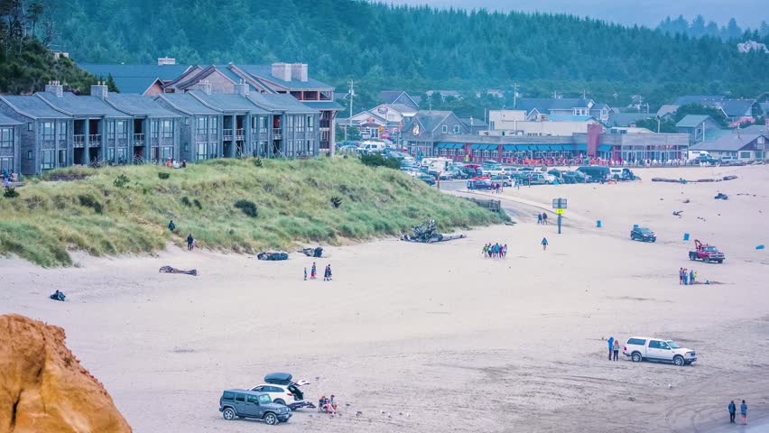 Scenic view of a coastal beach town in Oregon with buildings, sand dunes, and people enjoying the area. Vehicles are parked on the sandy beach.