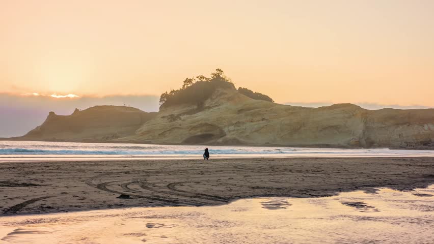 A person stands on the beach at low tide, watching the sunset over the iconic Haystack Rock formation at Cape Kiwanda State Natural Area in Pacific City, Oregon. The warm light reflects on the wet san