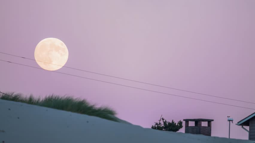A large, bright full moon rises in a soft pink and purple sky over sandy dunes and coastal vegetation near the Heceta Head Lighthouse in Coastal Oregon. The serene evening atmosphere captures the natu